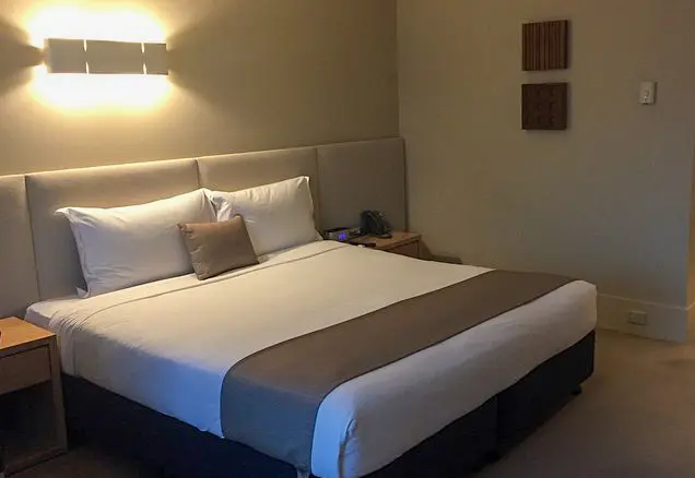 Modern hotel room at Crown Promenade Melbourne featuring a king-sized bed, large windows with city views, a work desk, armchair, and neutral-toned decor.