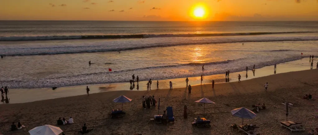 Sunset over Kuta Beach, Bali, with vibrant orange and pink hues reflecting on the ocean waves and silhouettes of people walking along the shoreline.