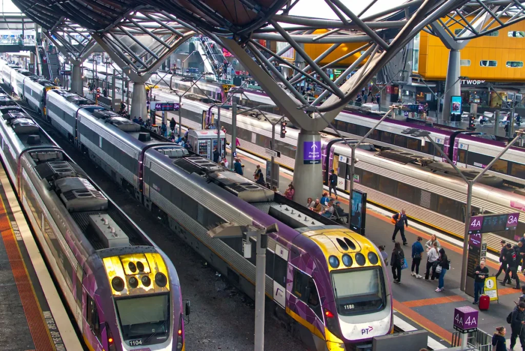 A busy scene at Southern Cross Station in Melbourne, showing multiple V/Line trains at the platforms, passengers walking and waiting, and the distinctive wavy architectural roof structure above.