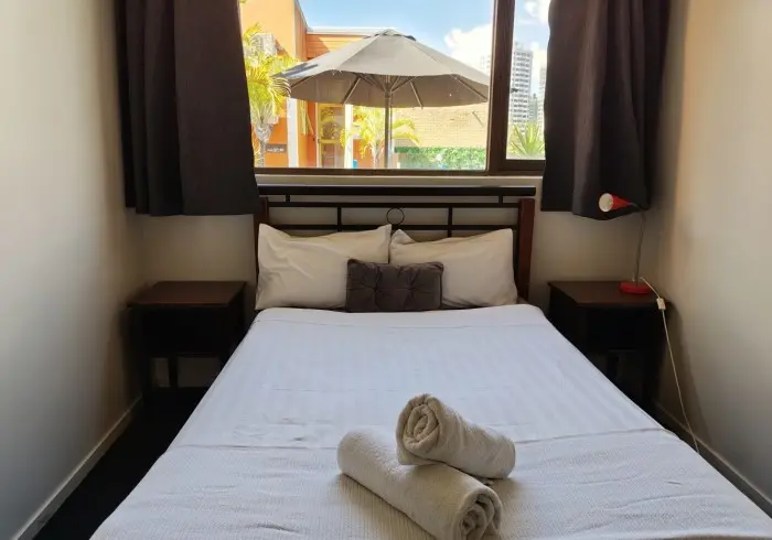 Small hotel room with a double bed made up in white linens, two rolled towels placed on top, and a window above the bed with black curtains partially open, revealing an outdoor umbrella and greenery.