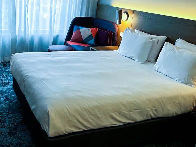Modern hotel room at Holiday Inn Sydney Airport featuring a large bed with white linens, a cushioned headboard with soft lighting, a work desk with a chair, a water bottle, and a cozy seating area with a colorful pillow near the window covered with sheer curtains.