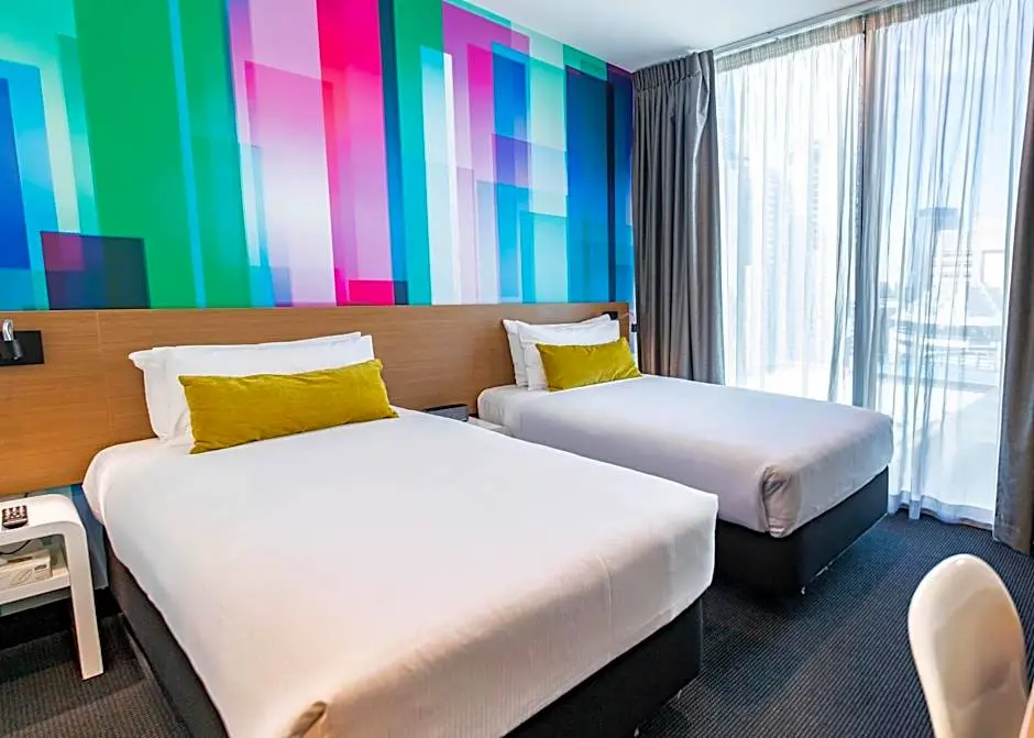 Modern hotel room with two single beds, each with white linens and a yellow accent pillow, set against a colorful geometric-patterned wall. Large floor-to-ceiling windows with sheer curtains allow natural light to fill the room.