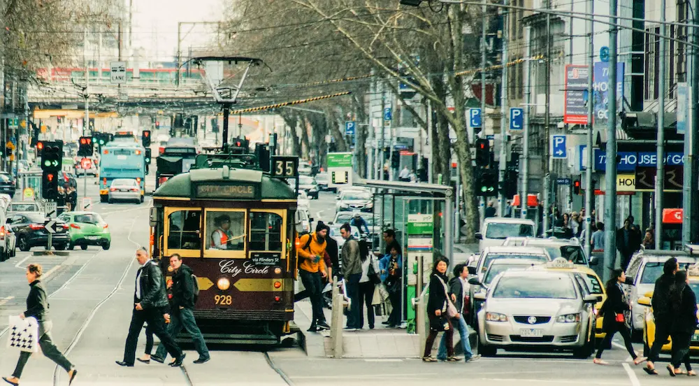 Busy Melbourne city street scene featuring a vintage City Circle tram, people crossing at a tram stop, and traffic including taxis and cars.