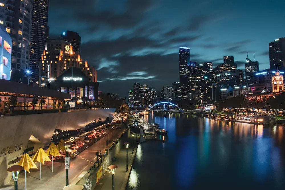 Nighttime view of Melbourne’s Southbank with city lights reflecting on the Yarra River, outdoor dining areas, and illuminated modern skyscrapers under a cloudy sky.
