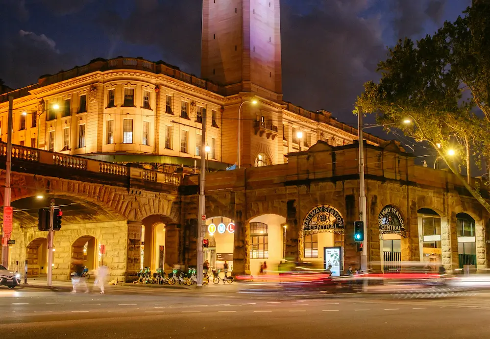 Central Station in Sydney illuminated at night, with bright lights highlighting the historic clock tower and modern buildings in the background.