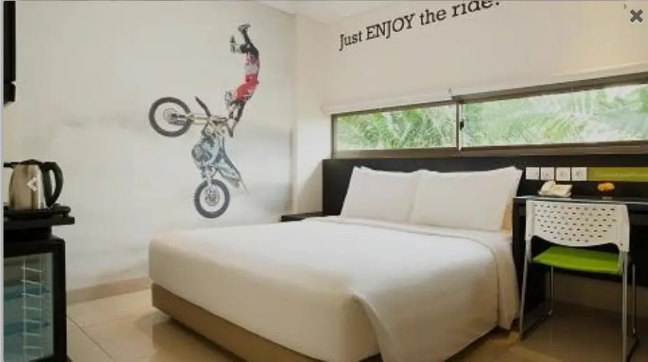 A bright hotel room at Grandma’s Seminyak with a queen bed, white linens, a wall mural of a motocross rider, a desk and chair, and a large window with green foliage outside.